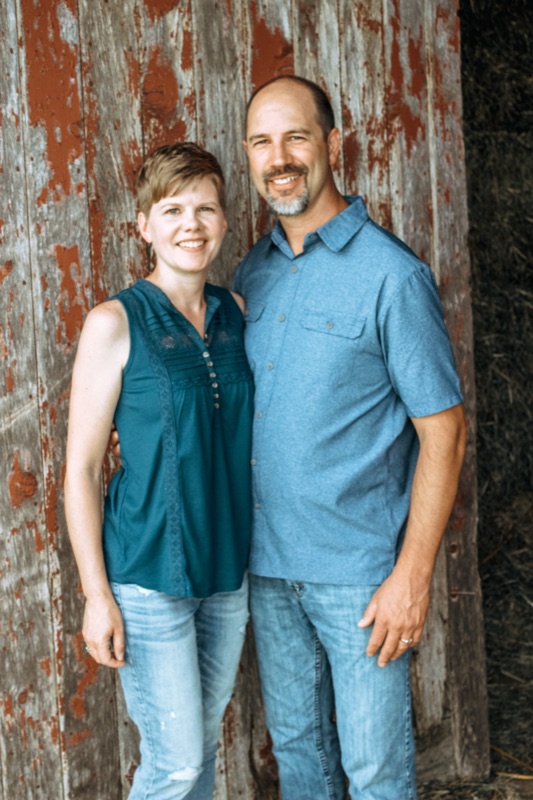 Kanon and Kathy Kulpa, founders of Barn & Bride, standing in front of a weathered red barn in Ellsworth, Wisconsin